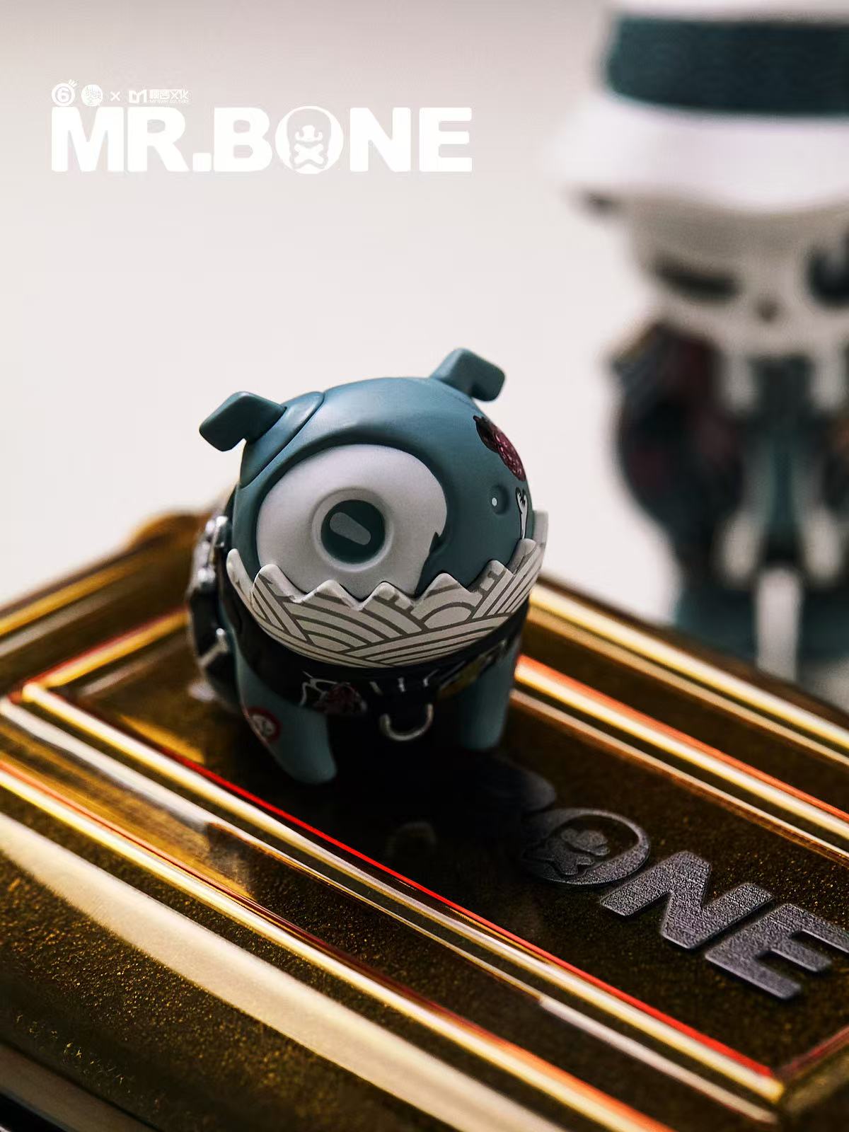Mr Bone Yakuza Limited Set 100% preorder features a slate gray designer vinyl toy with a bandana, on a gold-tone base, showcasing intricate textures.