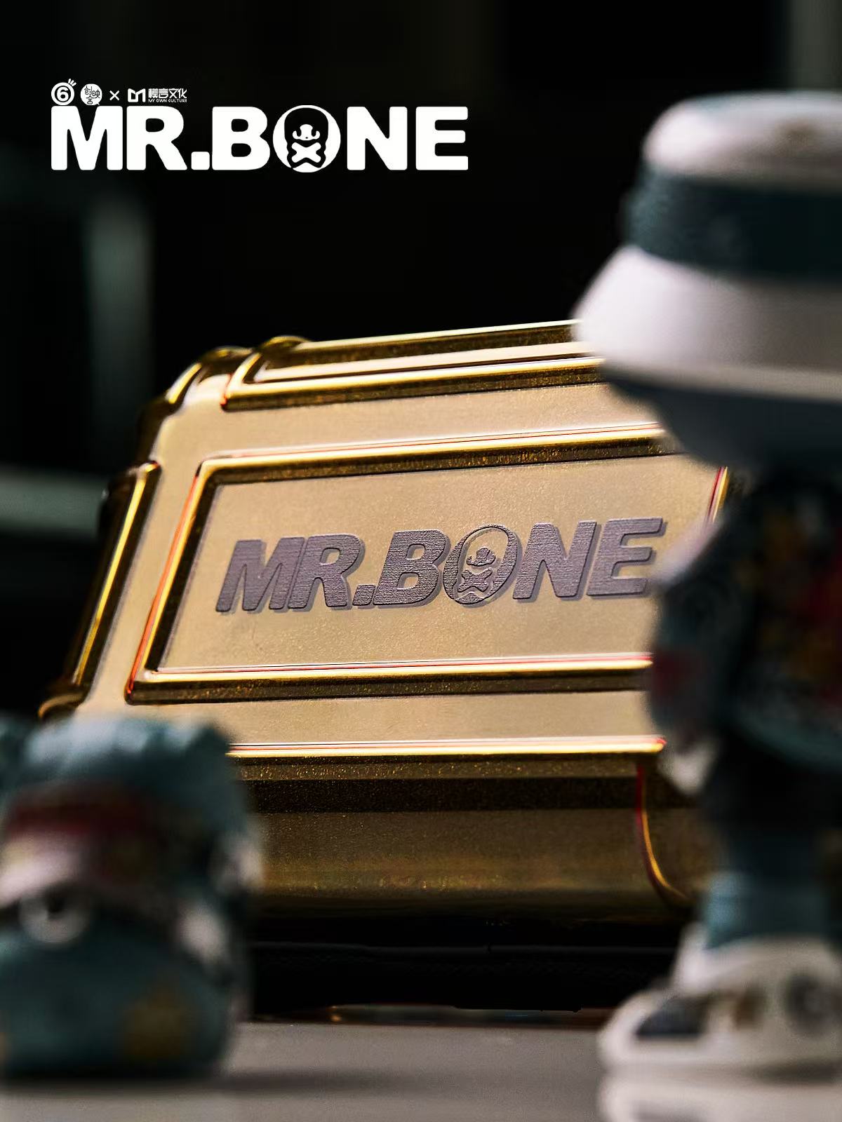Mr Bone Yakuza Limited Set 100% - Preorder features a gold case with embossed branding, including two out-of-focus collectible figurines in the foreground.