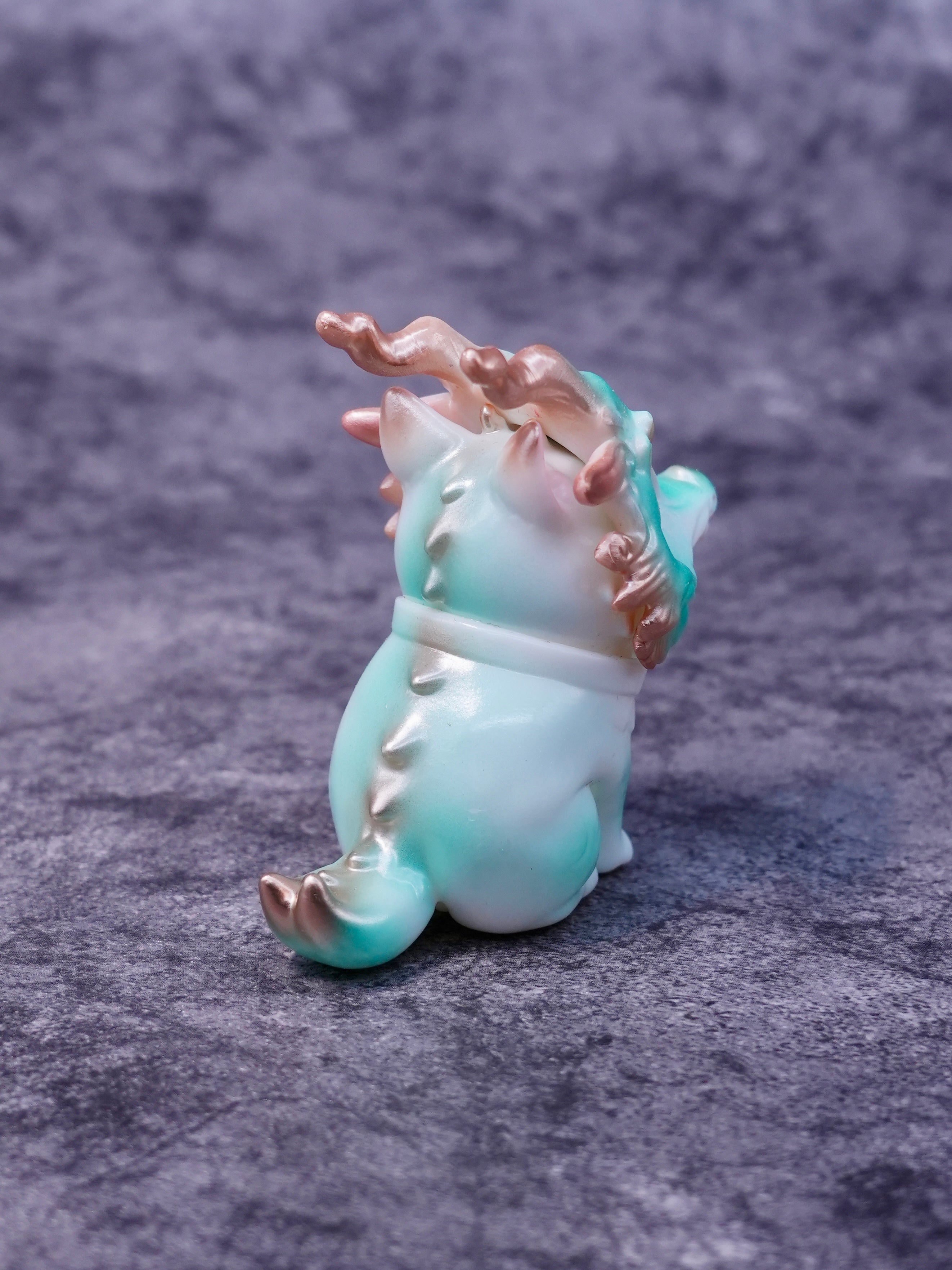 Little Azure Loong figurine, cat-dragon fusion with rose-gold horns, gradient body, and dorsal spines, viewed from behind on a gray surface.