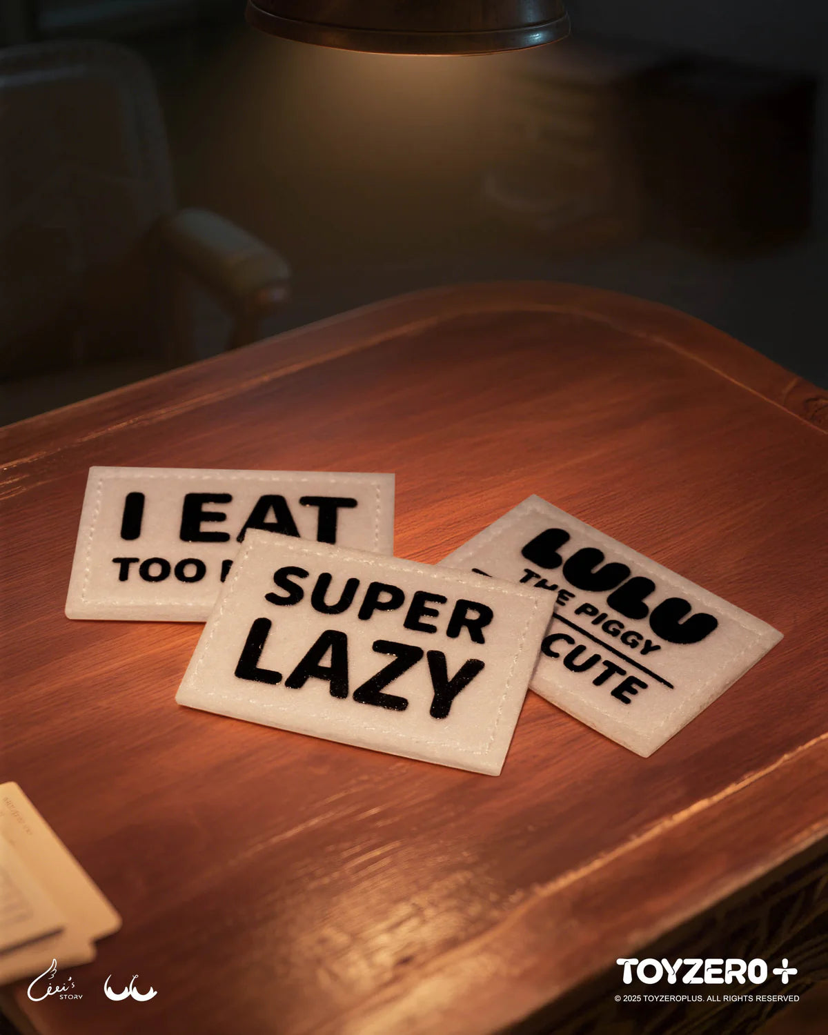 LuLu the Piggy LuLuLand keychain, featuring playful felt placards “SUPER LAZY” and “LULU THE PIGGY CUTE,” styled on a wooden tabletop. Preorder at Strangecat Toys.