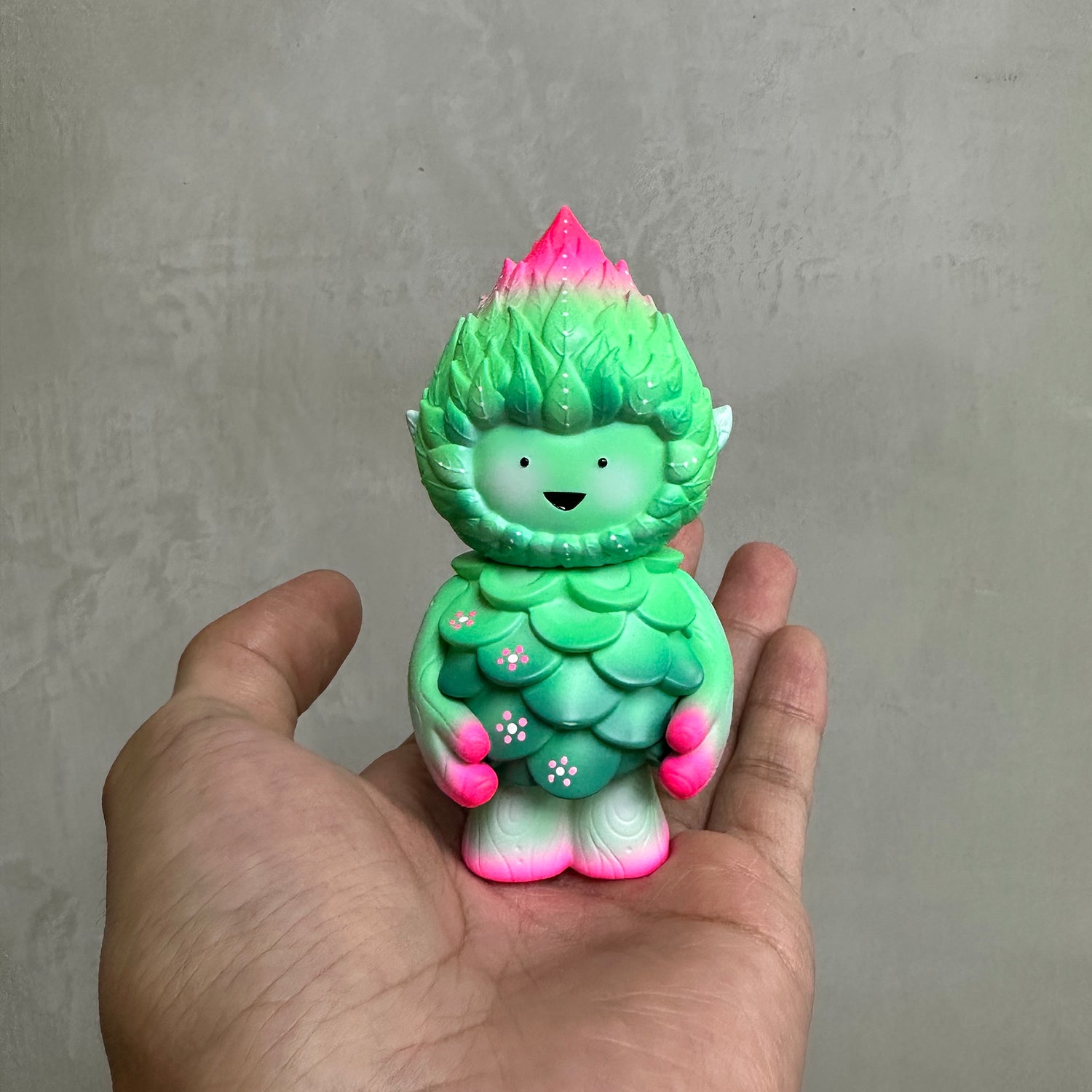 MUJUWORLD TREE SPIRIT SPRING EDITION vinyl figurine, leaf-covered, pink-tipped, smiling mask face, held in hand; preorder from Strangecat Toys, 4 inches tall.