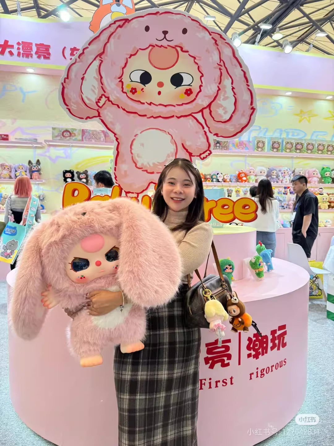 Woman holding oversized Baby Three AI 800% stuffed animal, part of a preorder series, measuring 19.7-21.7 inches, from Strangecat Toys. Ships late July 2025.