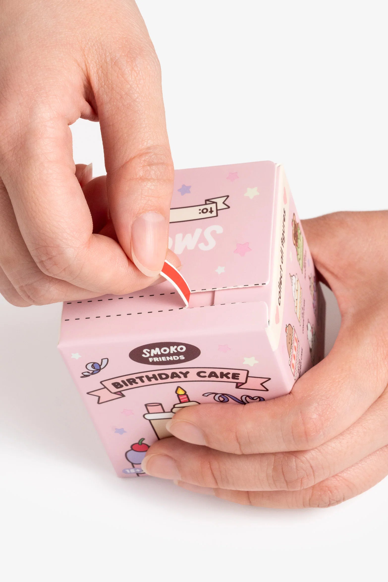 Hands opening a pastel-pink Smoko Friends Birthday Cake Blind Box Figure with festive designs, revealing a surprise collectible toy inside.