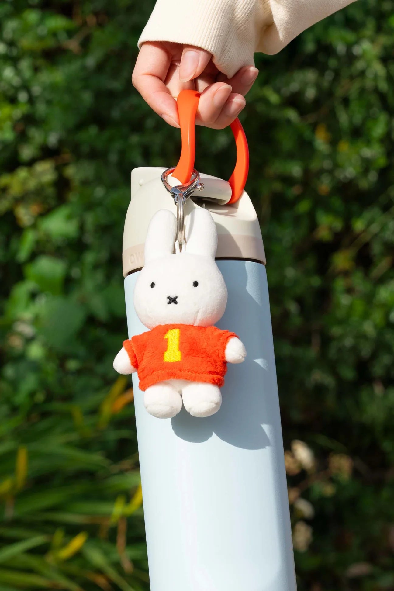 Pale blue water bottle with orange loop, featuring a Smoko Miffy #1 Bag Charm, a plush bunny in an orange sweater, outdoors.