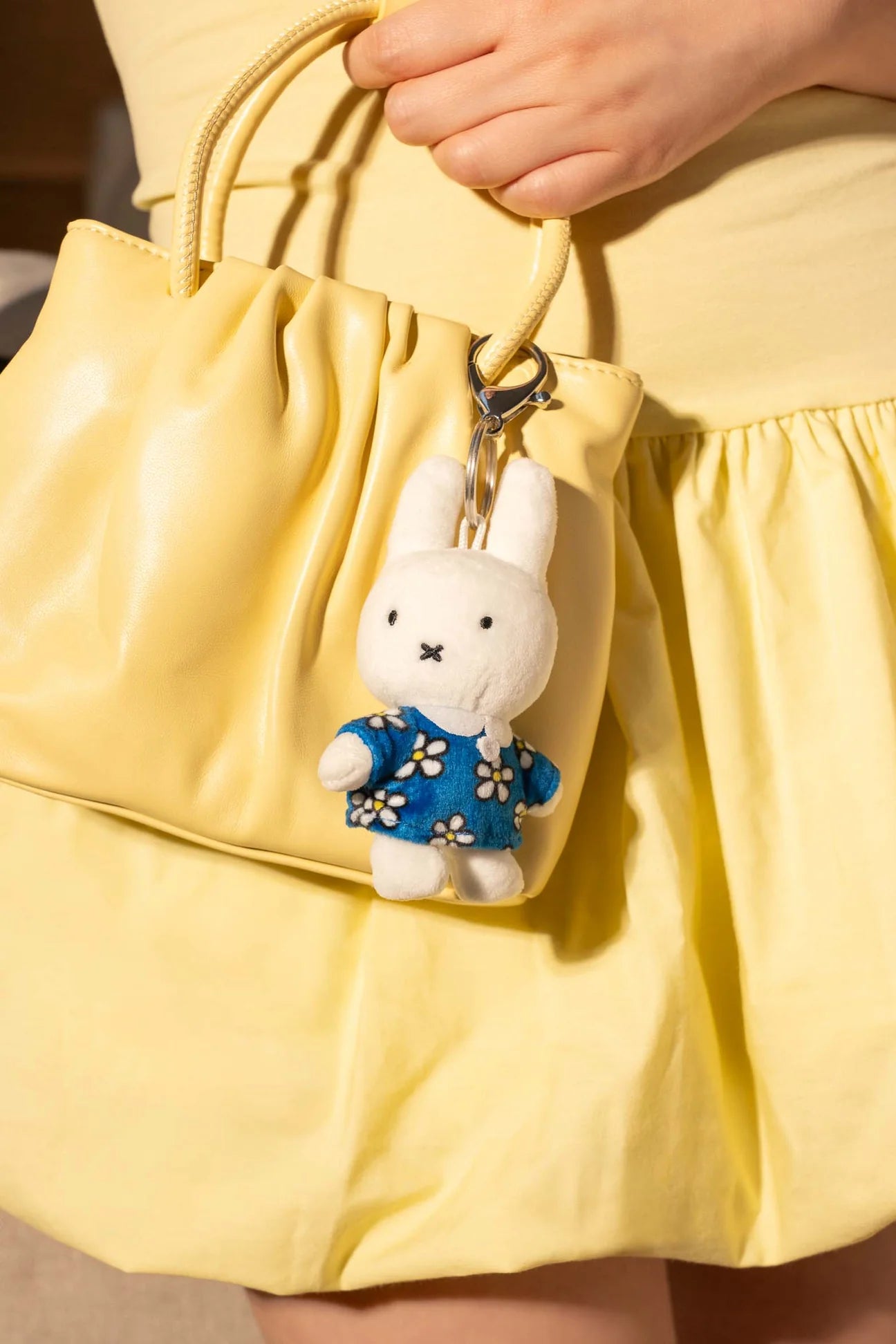 Smoko Miffy Blue Daisy Bag Charm attached to a soft yellow handbag, featuring Miffy in a blue daisy-patterned shirt, held by a person in yellow attire.