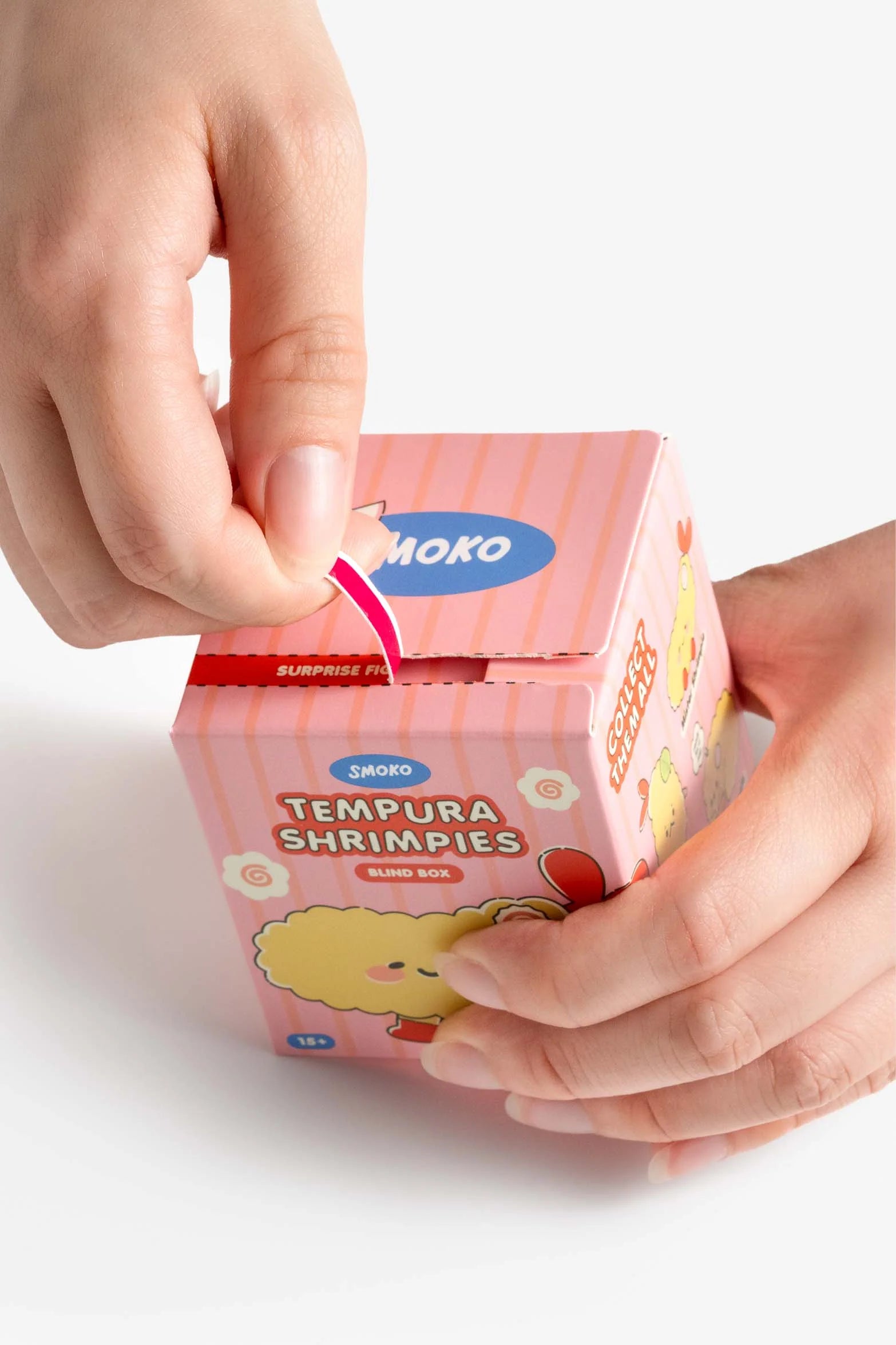 Hands opening a Smoko Tempura Shrimpie Blind Box, revealing a surprise collectible figure inside. The box features a cute tempura shrimp character.