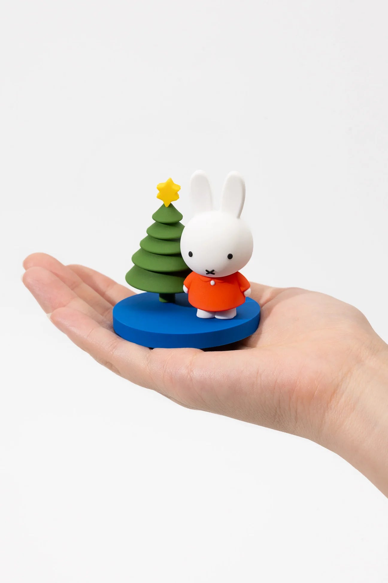 Hand holds a Miffy in the Snow Blind Box Figure featuring a white rabbit with an orange dress beside a tiered Christmas tree on a blue base.