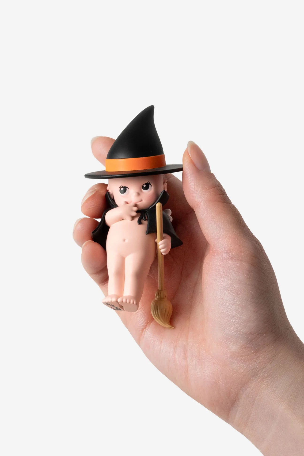Smoko Kewpie Halloween Blind Box Figure: A hand holds a resin figurine of a baby witch in black cape and hat, clutching a tiny broomstick.