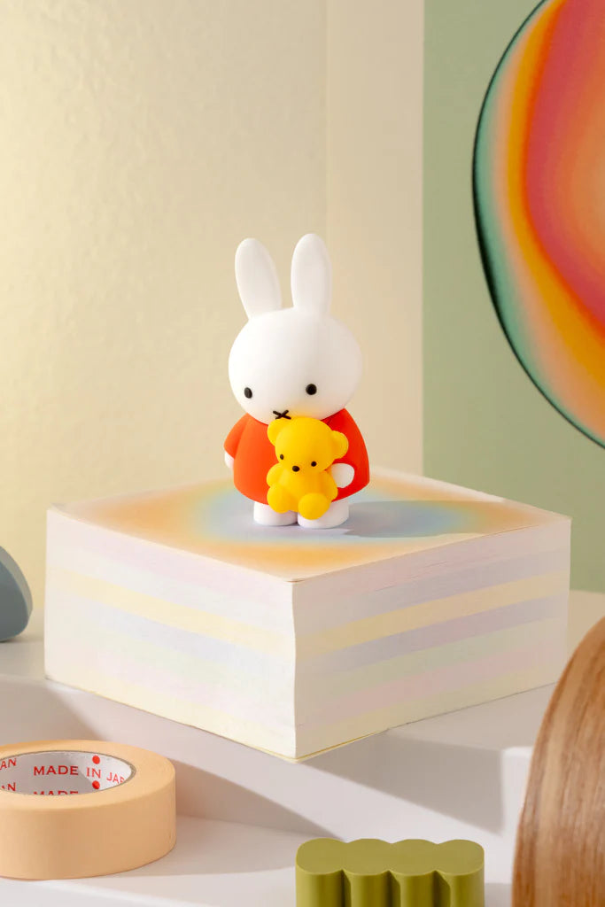 Miffy Doing Things Blind Box: White rabbit figurine in red outfit holding a yellow teddy bear, standing on pastel-striped notes with tape and olive object nearby.