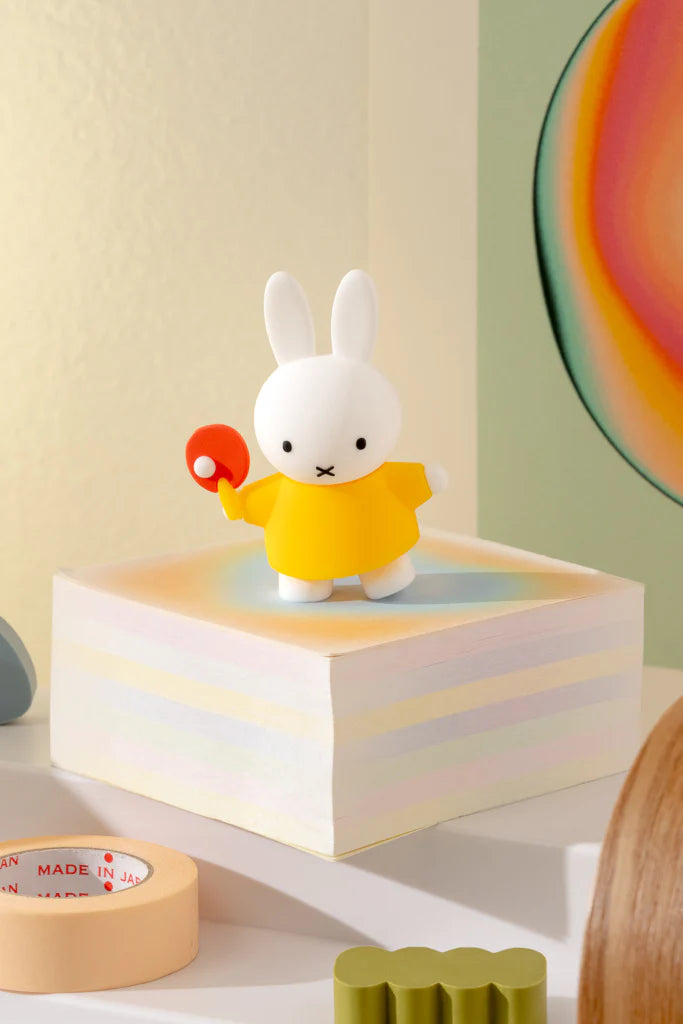 Miffy Doing Things Blind Box: A white rabbit figurine in a yellow tunic holding a red paddle, displayed on a pastel-striped block, part of a collectible vinyl series.