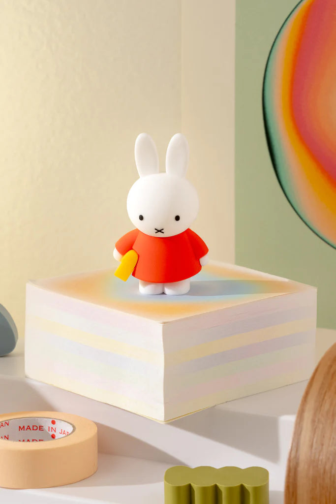 Miffy Doing Things Blind Box: Vinyl toy of Miffy in a red dress holding a yellow item, standing on pastel-striped notepaper with masking tape nearby.