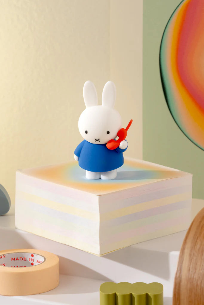 Miffy Doing Things Blind Box: Miffy figurine in blue with red phone, atop a notepad, surrounded by desk items. Collectible, surprise figure, officially licensed.