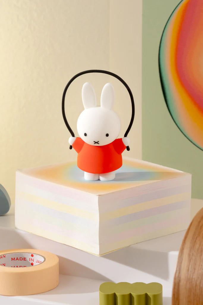 Miffy Doing Things Blind Box: A white rabbit figure in a red-orange dress, holding a jump rope, stands on pastel memo pads with minimal desk props nearby.