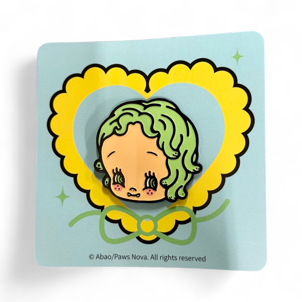 Baby Medusa Metal Badge featuring a cartoon head with wavy green hair, big lashes, rosy cheeks, and tiny fangs on a decorative backing card.