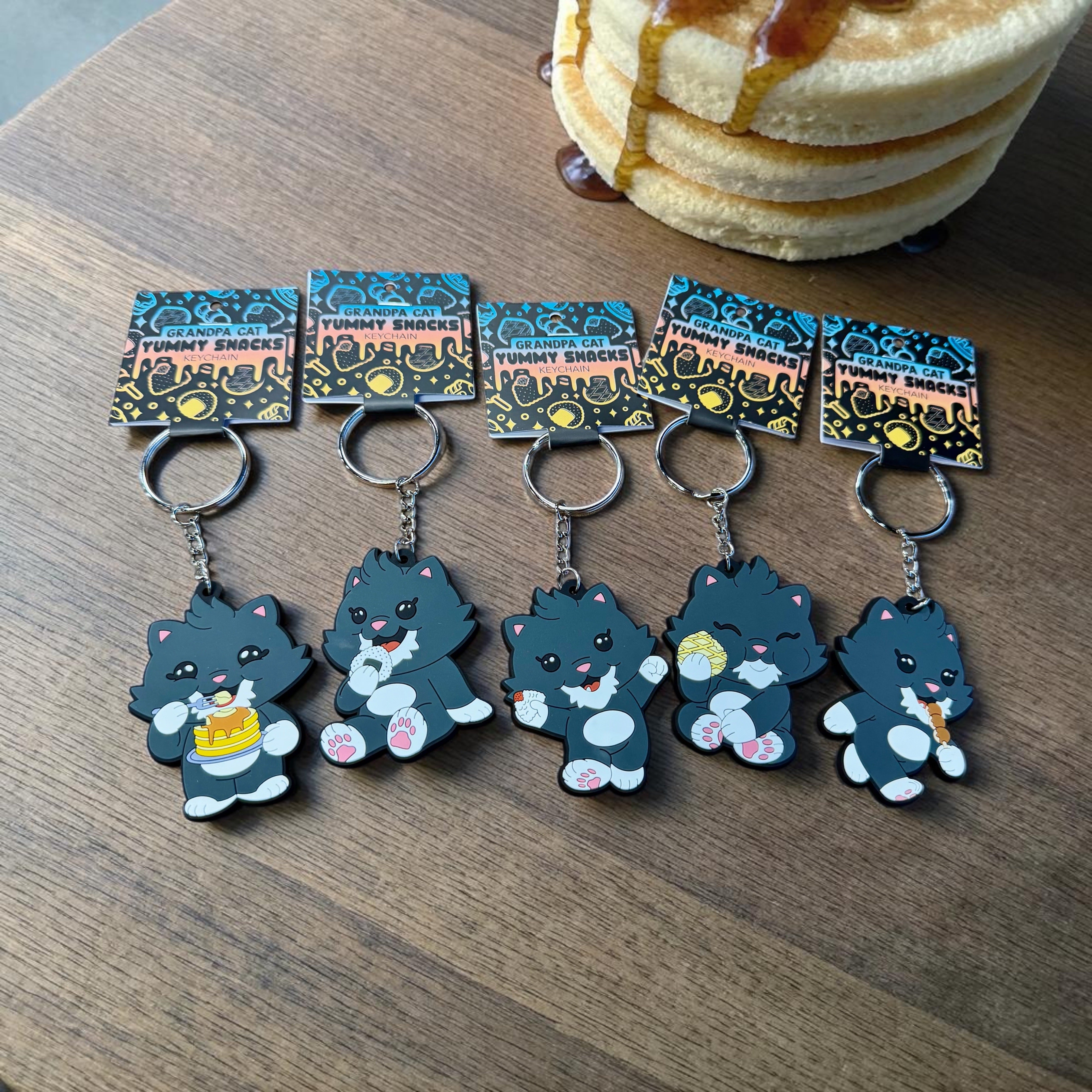 Grandpa Keychain - Japanese Snack Series features five silicone keychains of cartoon cats with snacks, displayed on a wooden surface.