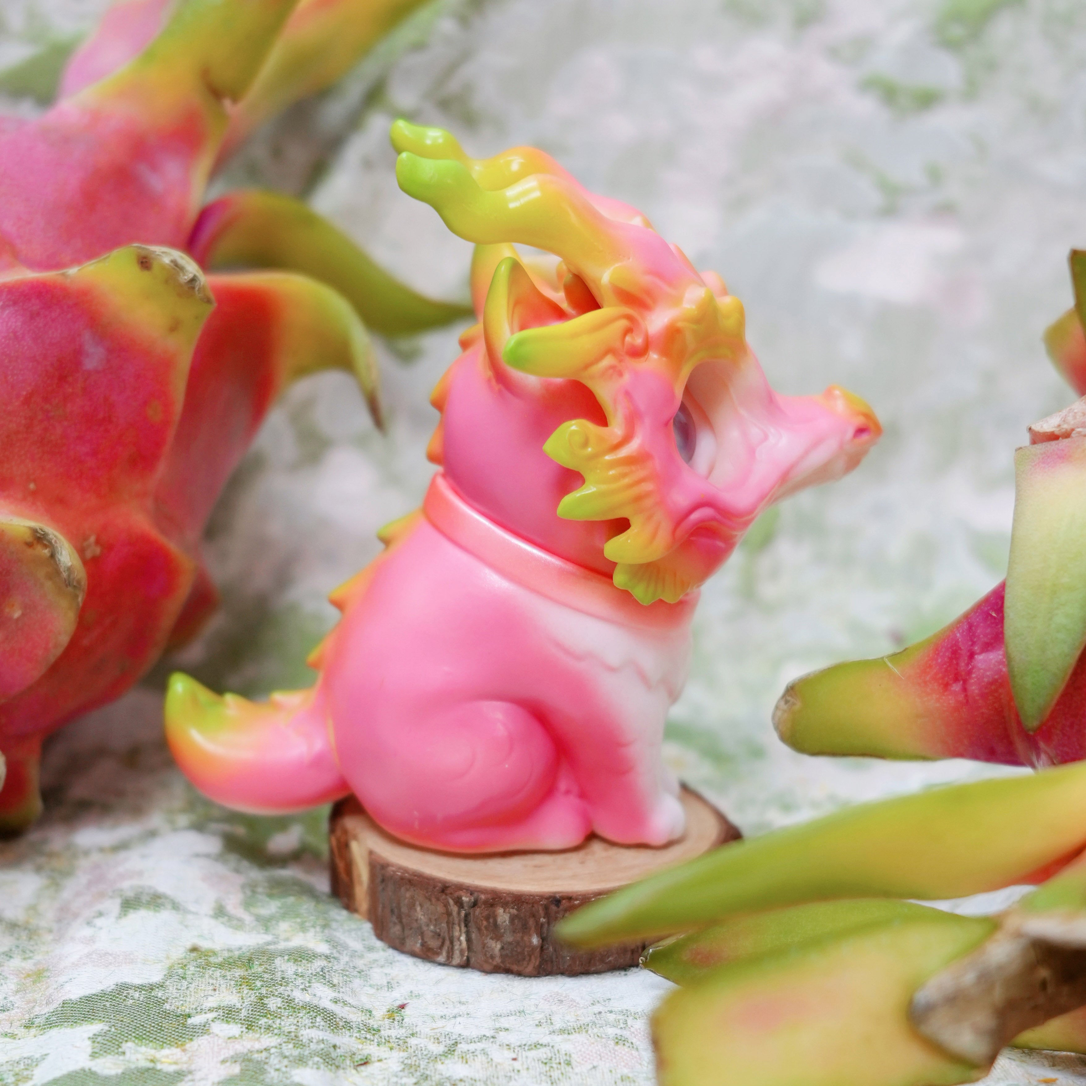 Pitaya Loong cat by HOTRU: A pink and lime green dragon-dog figure with curling horns and frills, on wood, surrounded by dragon fruit.