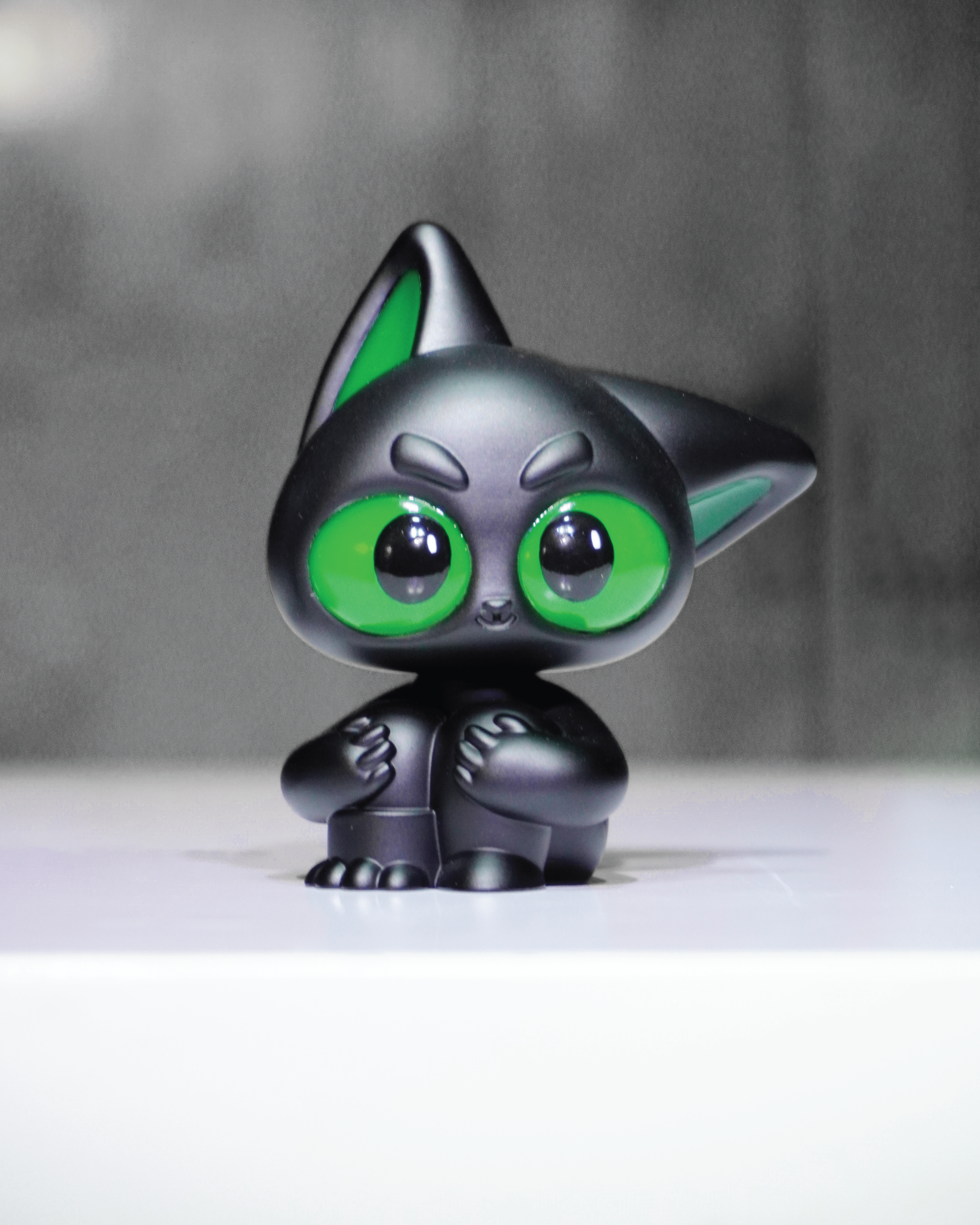 Noir by Wapu: A 3 black resin figurine of a cartoonish creature with bright green eyes, sitting upright on a white surface.