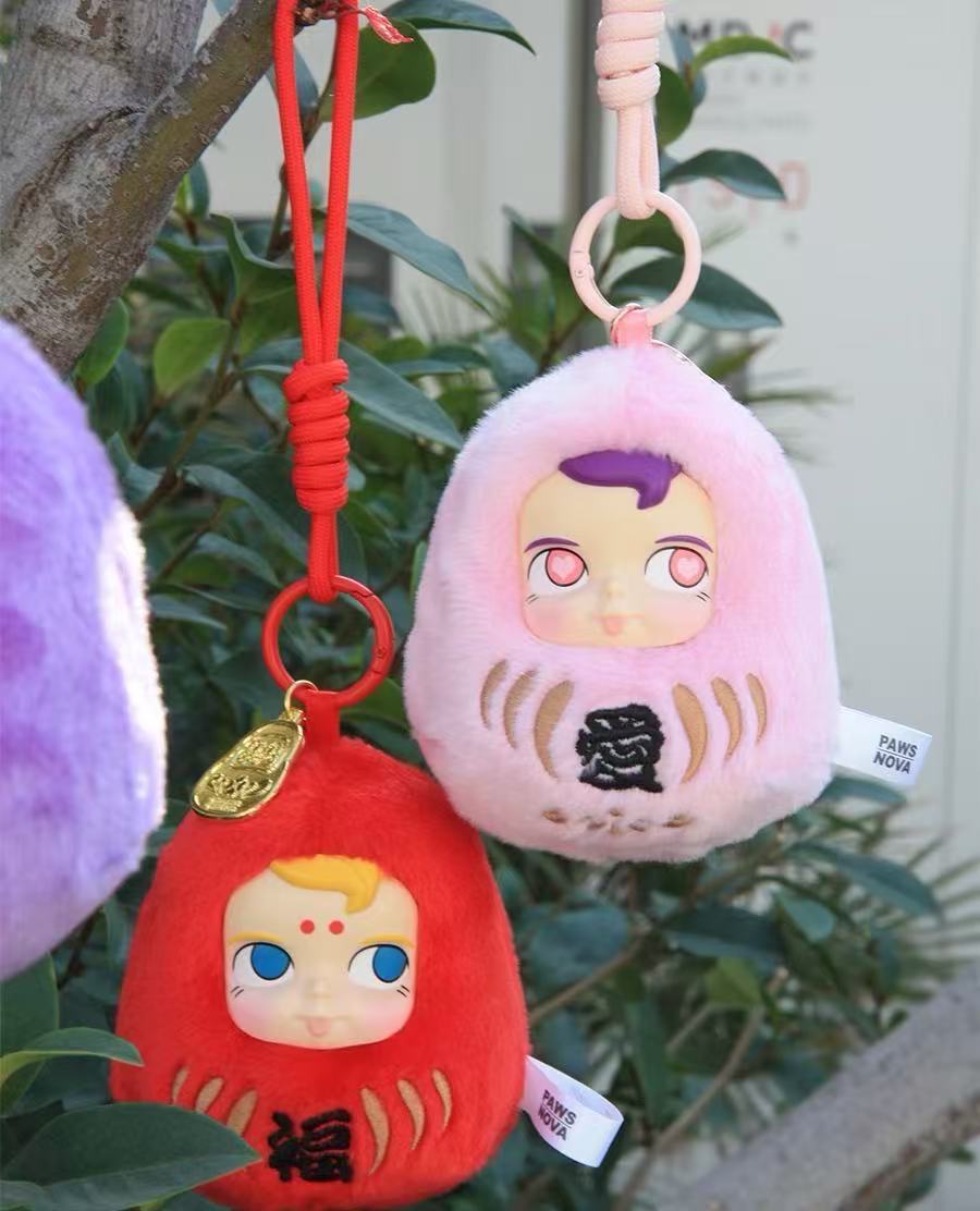DHARMA SAMSAM VINYL FACE PLUSH PENDANT BLIND BOX charms with cartoon-baby faces hanging from a branch, featuring embroidered Japanese characters and PAWS NOVA tags.