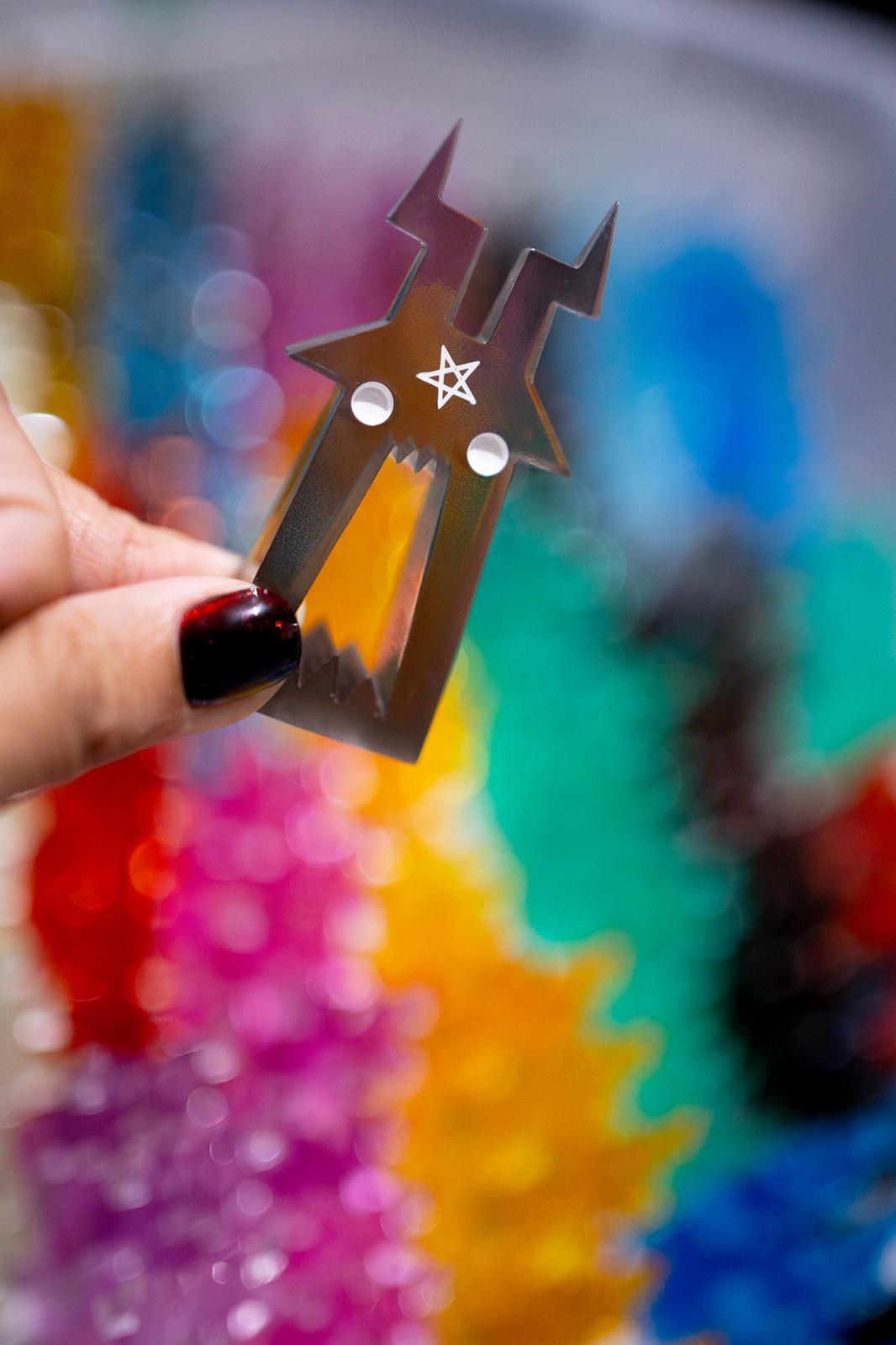 Hand with red-tipped nails holds a small metal 'Phillips' monster figure from Zintears, featuring lightning-bolt horns and a pentagram, against a colorful backdrop.