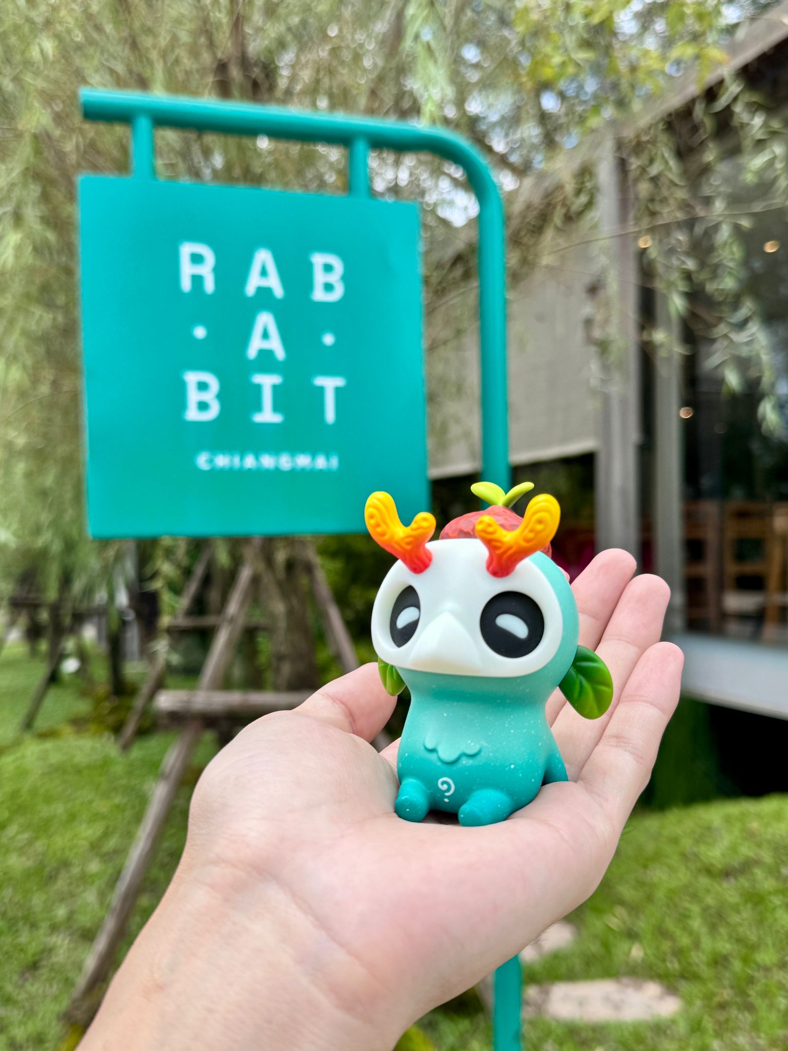 Ori muffinn by Madkids: A small, mint-green vinyl toy with leaf-shaped ears and orange antlers, held in hand against a leafy café backdrop.
