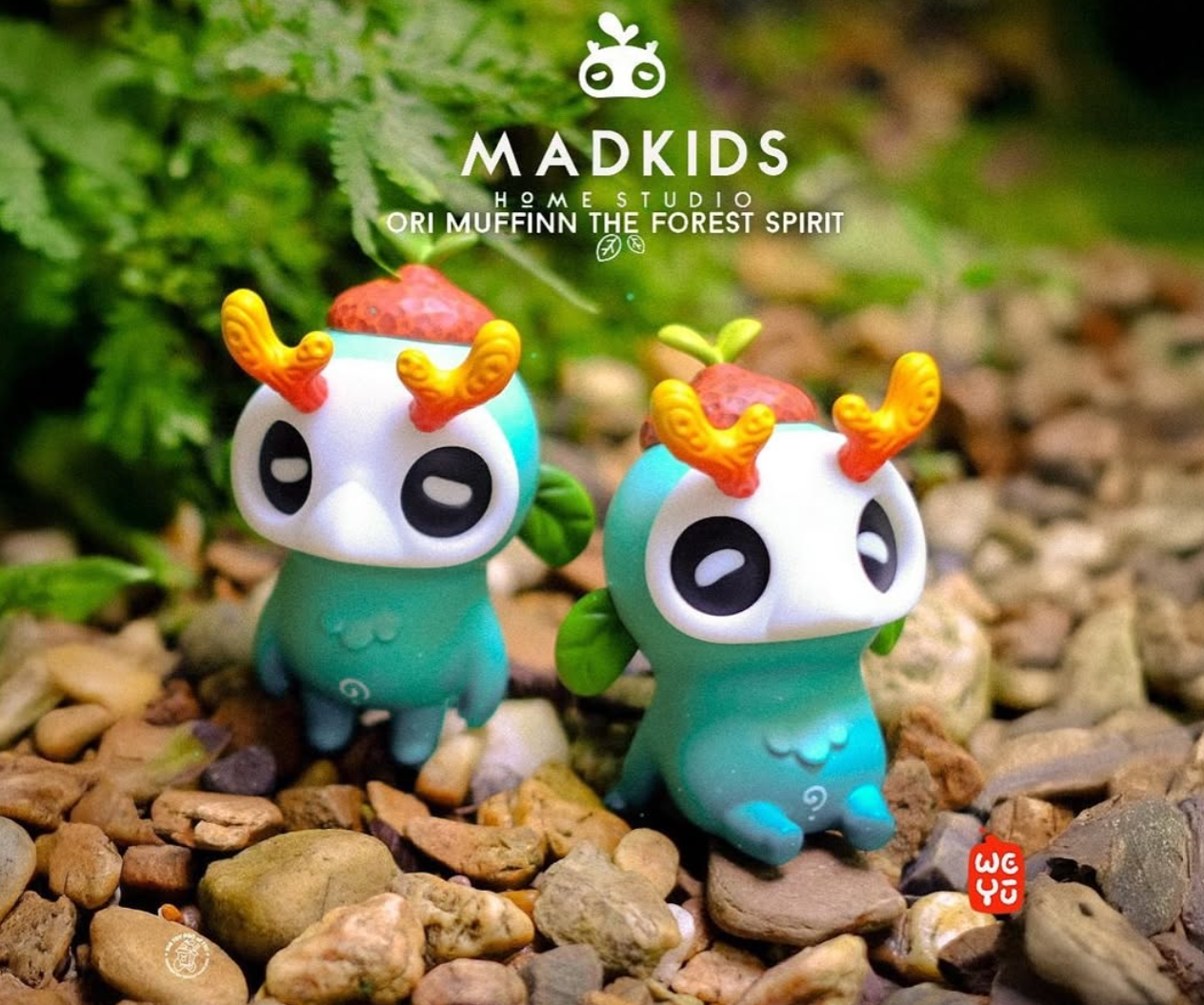 Ori Muffinn by MADKIDS: Mint-green collectible figures with white mask-like faces, acorn caps, antler-like horns, leaf ears, and spiral bellies, set on pebbles.