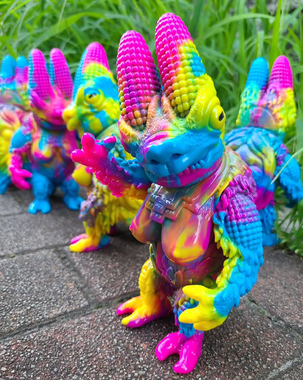 Pachnadorah by Cesar Zanardi: Colorful vinyl figures with horned heads and scaly limbs, showcasing unique paint patterns. Set on a paved surface with grass background.