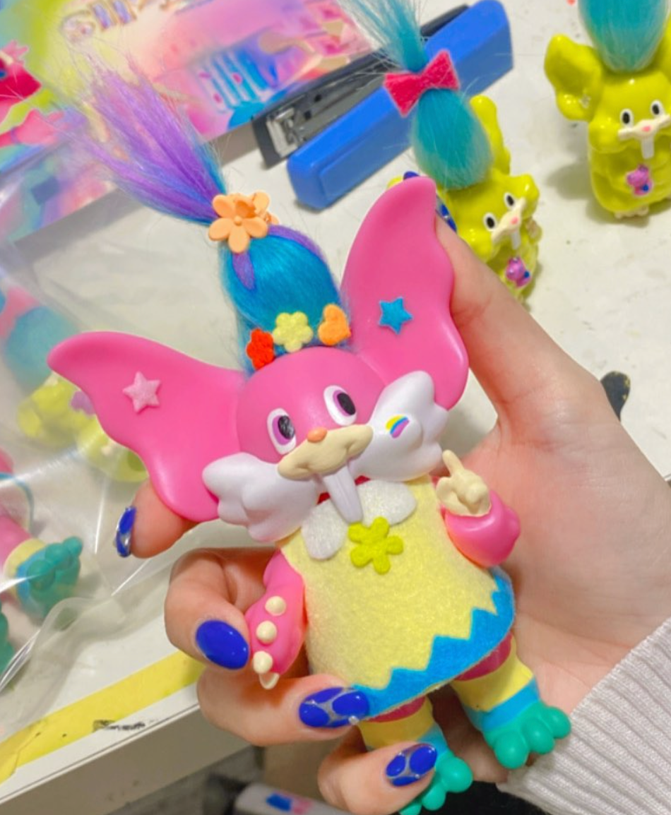 Close-up of Trolls Trolls by Vivian Wang, a 13cm vinyl art toy with oversized ears, rainbow cheek decal, fuzzy yellow dress, and vibrant tufted hair.