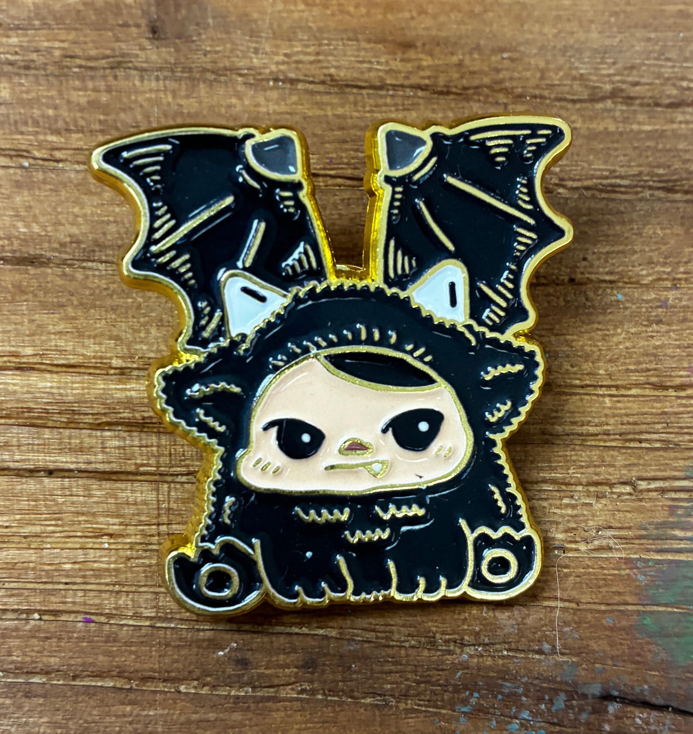 Bloom Pins by Pucky: A chibi-style enamel pin of a character in a black costume with bat wings and cat-ear tufts, displayed on a wooden table.