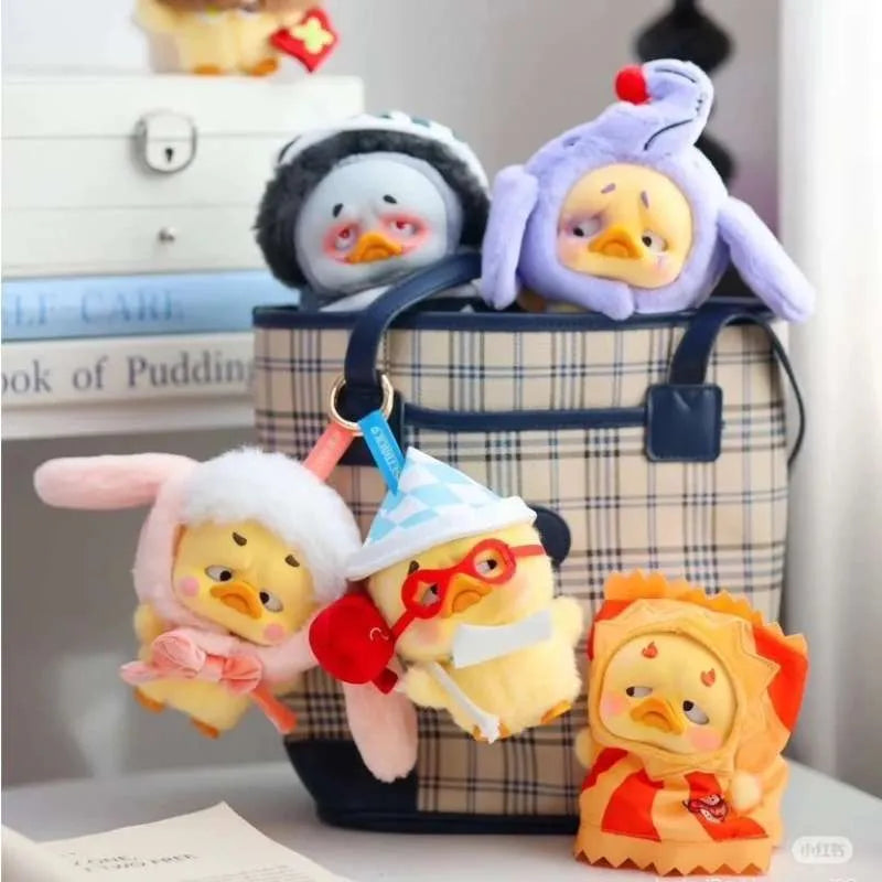 Plaid tote with five Upset Duck - Pocket Crazy Circus Duck keychain toys, each in distinct costumes, hanging on a table with Book of Pudding behind.