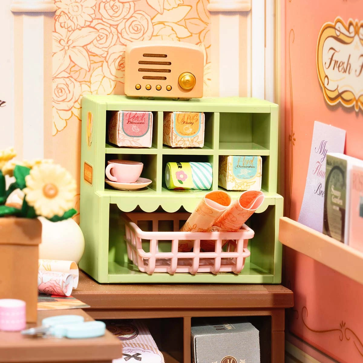 DW017B Fresh Flower Shop Rolife DIY Miniature House: A pastel-toned tabletop vignette featuring decorative boxes, a pink teacup, and a retro radio, embodying cozy craft shop charm.