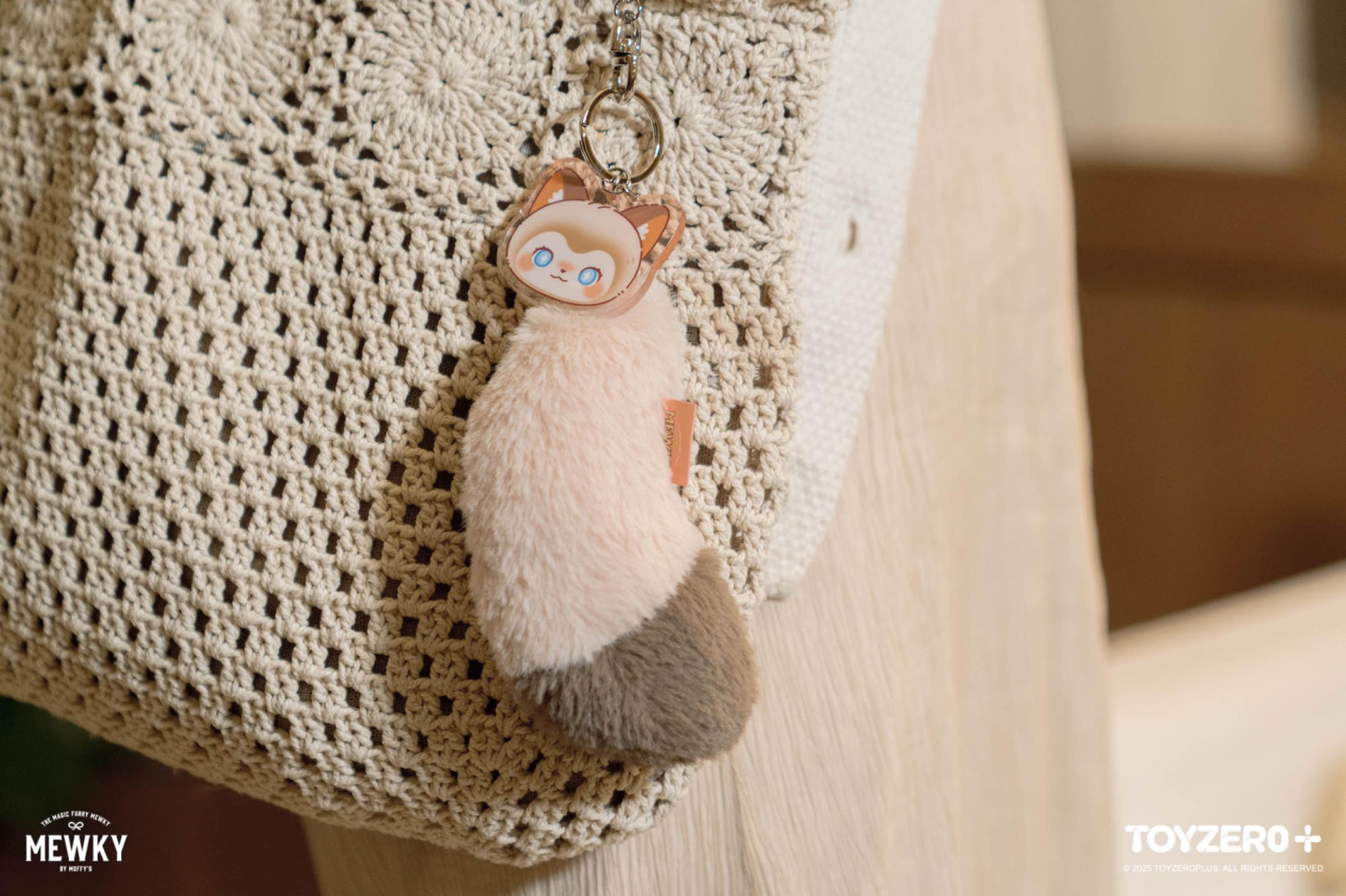 Moffy's Mewky Cat Tail Keychain - Siamese Cat: A plush pink-and-grey tail keychain on a cream-crocheted tote, featuring a cartoon animal face with blue eyes.