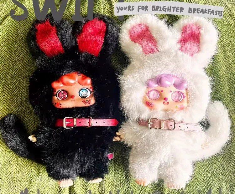 Migo Twins 400% plush dolls in black and white animal suits with big eyes and rosy cheeks, posed on a green fabric backdrop.