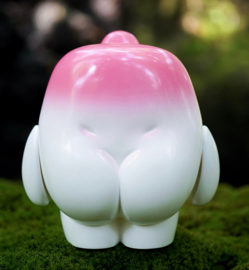 100% MALLOW Pink by MUPA, a small, smooth vinyl art-toy figure, stands on moss. Its glossy pink top transitions to a white lower half.