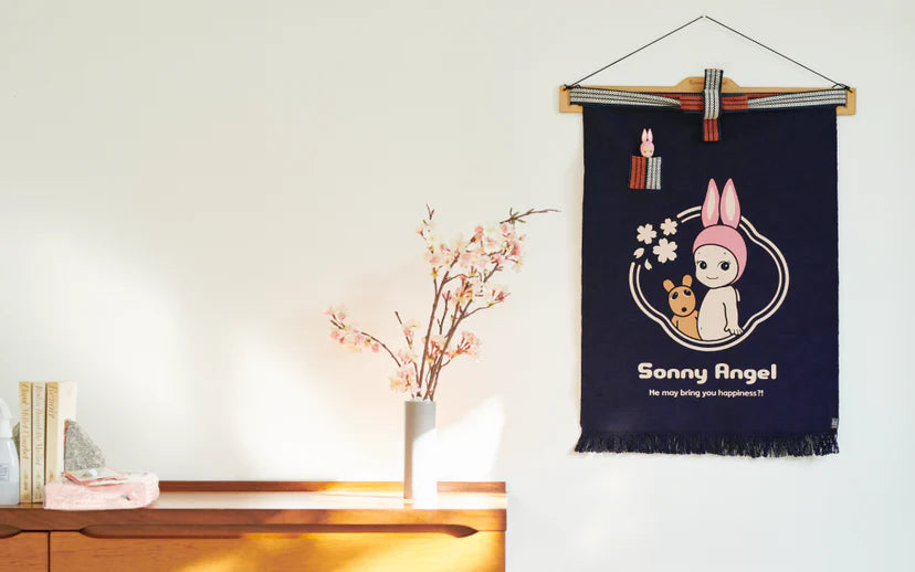 Sonny Angel x Anything Maekake Apron displayed with a navy fabric banner, books, a pink cloth, a decorative stone, and a vase with cherry blossoms.