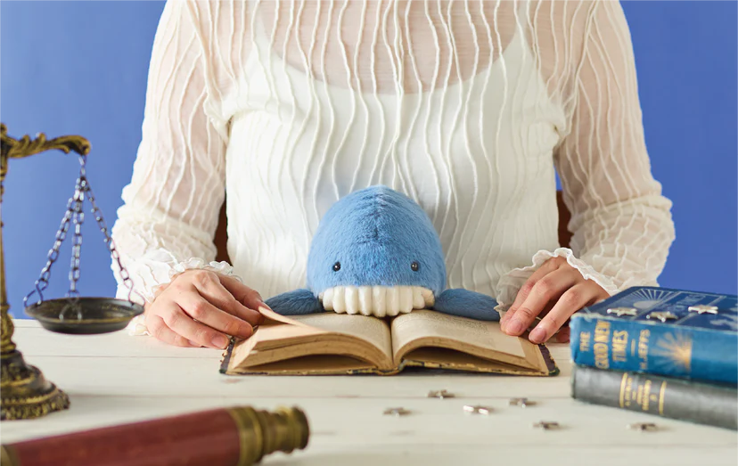 Posture Pal - Marine: A blue stuffed-whale toy with white teeth sits on an open book, surrounded by scales, books, puzzle pieces, and a telescope.