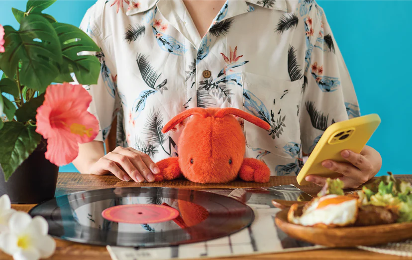Person at table with tropical shirt holds yellow phone, surrounded by a plush orange bunny, vinyl record, hibiscus plant, and breakfast.