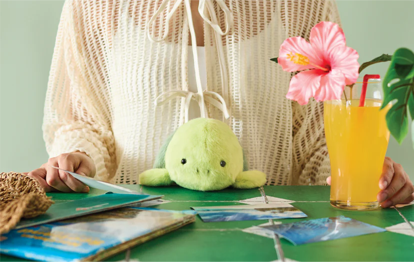 Turtle-themed Posture Pal amidst travel brochures, straw hat, and a hibiscus-garnished juice, embodying a relaxed, vacation vibe for ergonomic desk comfort.