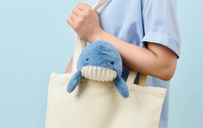 Person holding a canvas tote with a Posture Pal - Marine Whale plush, designed to support posture at desks, peeking out, showcasing its playful utility.