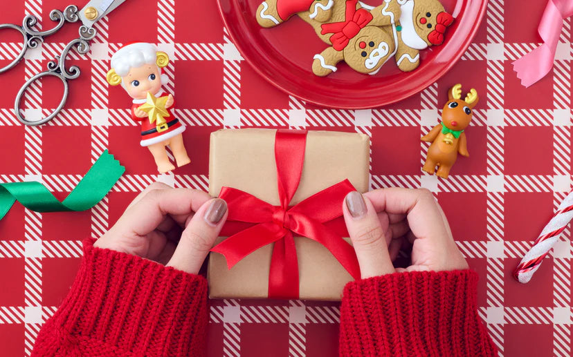 Santa's Little Helper Series toy doll in a festive gift-wrapping scene, surrounded by gingerbread cookies, a reindeer figurine, and holiday ribbons.