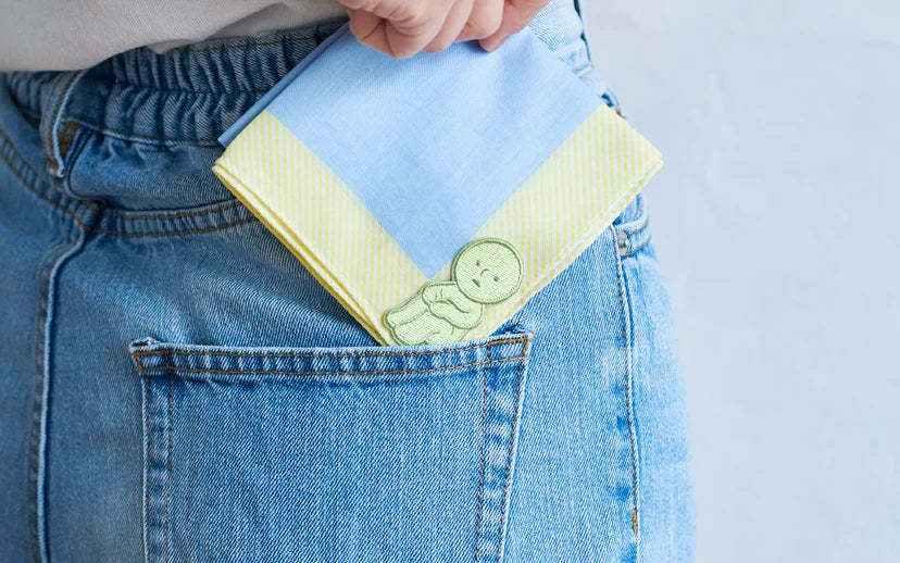 Hand tucks a two-toned SMISKI Embroidery Sticker featuring a cute green character into the back pocket of light-blue denim jeans.