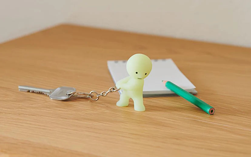 Smiski Keychain on a wooden surface next to a green pencil, showcasing a small glowing figure designed to help locate keys.