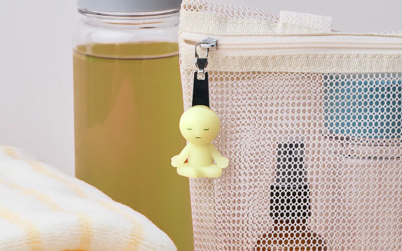 SMISKI ZIPPERBITE - Vol. 2 clipped to a mesh travel pouch with toiletries, featuring a meditating figurine that glows softly in the dark.