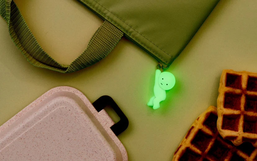 Smiski ZipperBites keychain attached to a green bag, placed next to a waffle. The keychain glows in the dark.