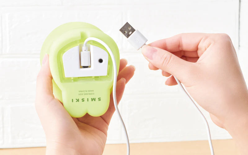 A hand holding a Smiski Touch Light, featuring Smiski daydreaming with a Smiskitty on its head. Tap to adjust brightness levels. Includes micro USB charger. Dimensions: W83 x D92 x H173mm.
