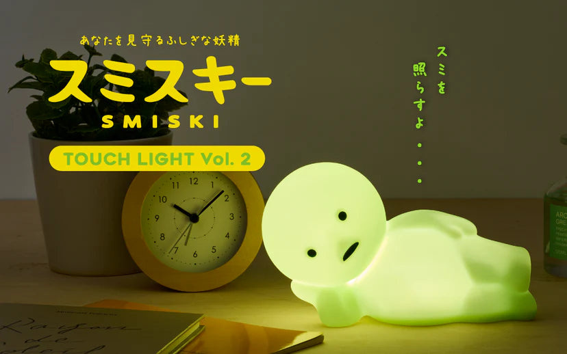 SMISKI Touch Light Vol. 2: A glowing green figure on a wooden surface, next to an alarm clock, plant, notebooks, and bottle, offering adjustable lighting.