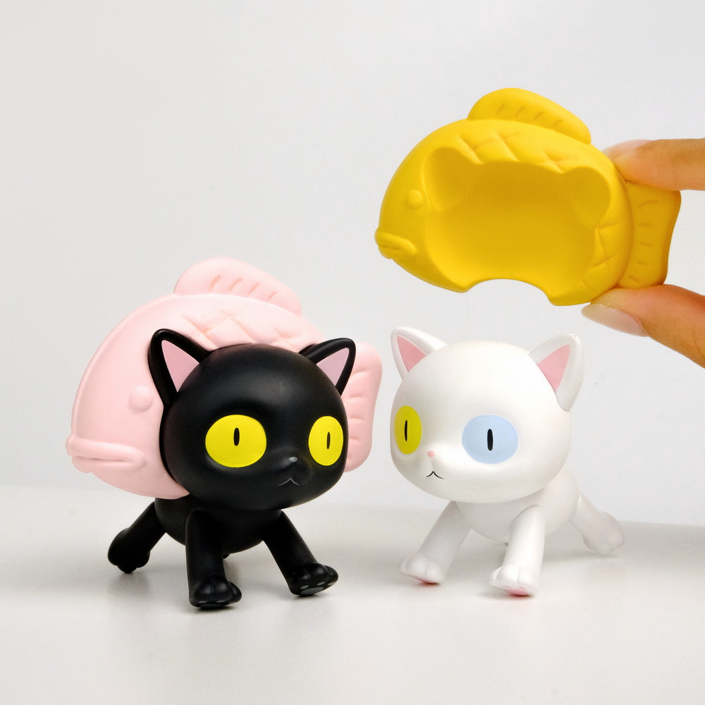 Two cat figurines from Gacha Bad Cats Blindbox, one black with pink shell, one white. Hand holding yellow shell, showcasing collectible toy designs.