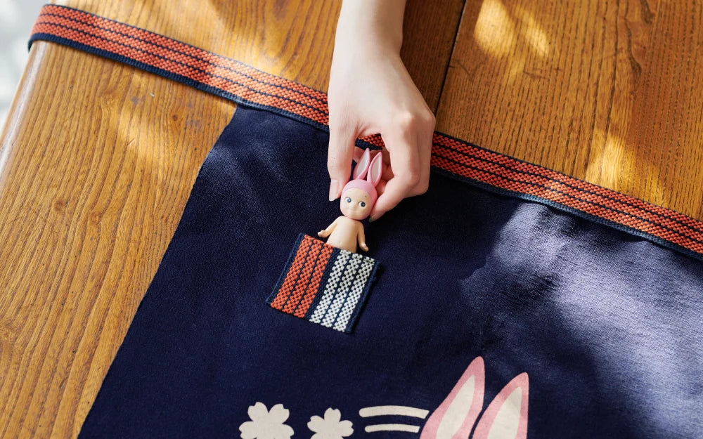 Hand holding a Sonny Angel x Anything Maekake Apron, showcasing a mini figure pocket feature, highlighting the collaboration's unique blend of tradition and art toy style.