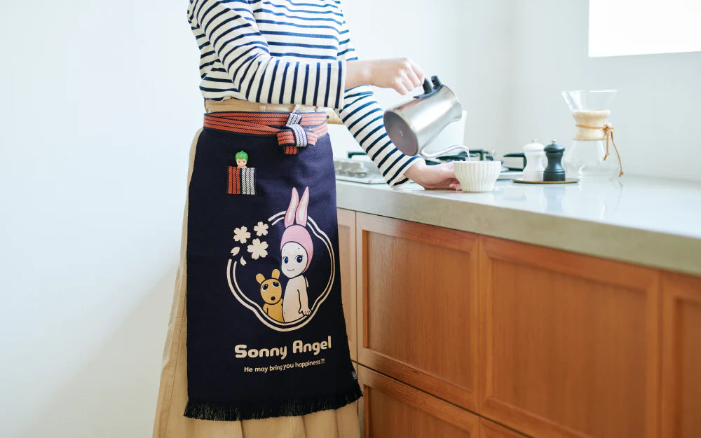 Person pouring from gooseneck kettle into cup, wearing Sonny Angel x Anything Maekake Apron, featuring cartoon designs, with Chemex and salt and pepper mills nearby.
