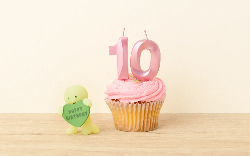 SMISKI Birthday Series: Vanilla cupcake with pink frosting, 10 candles, and a lime-green SMISKI figurine holding a HAPPY BIRTHDAY sign on a wooden surface.