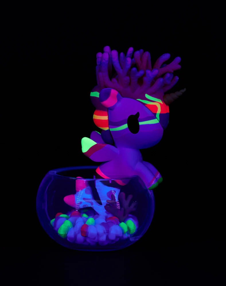 Neon Reef Mermicorno Blind Box figurine glows under UV light, perched on a glass bowl filled with luminous pebbles and coral-shaped elements.